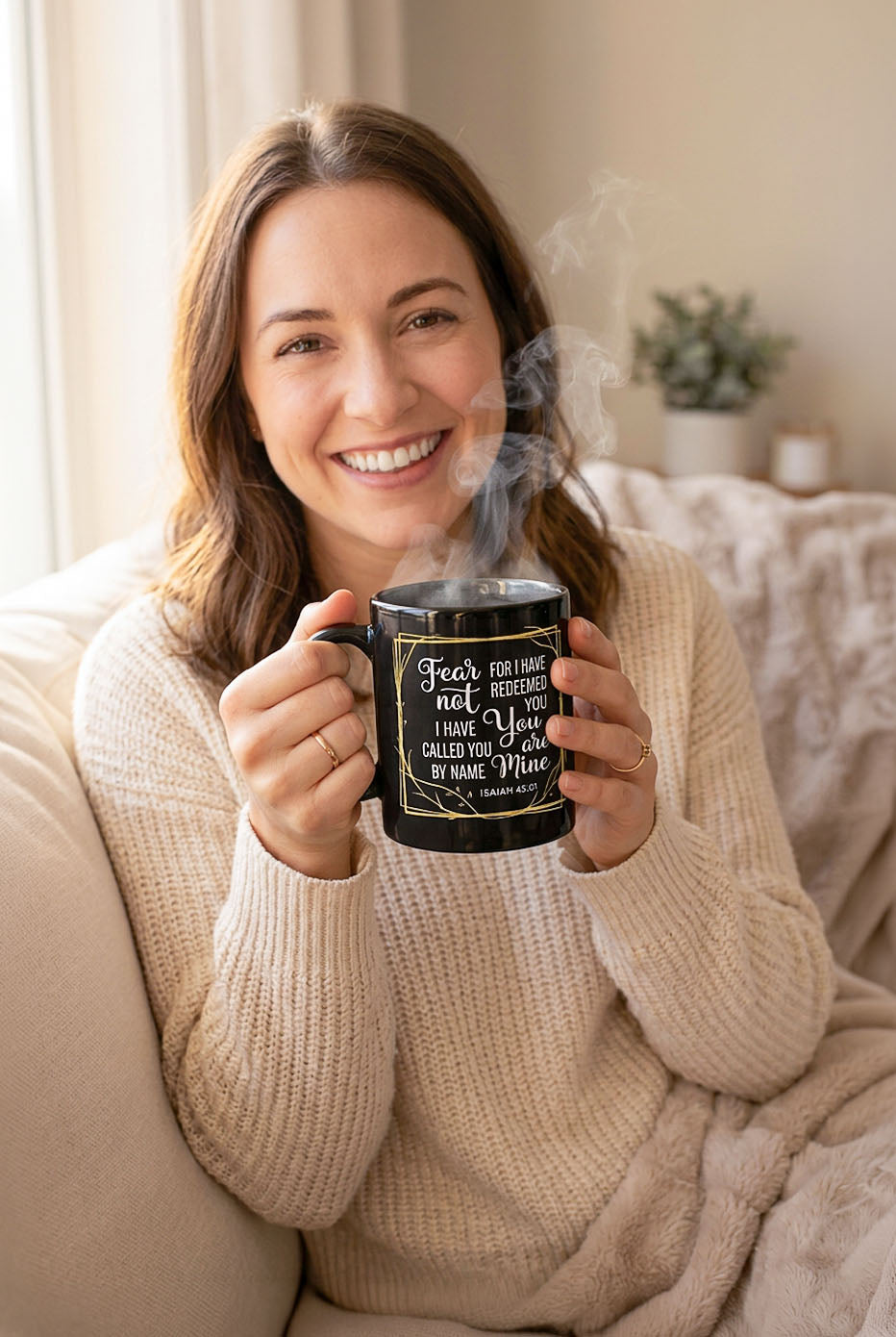 Smiling woman in a cream sweater holding a steaming black Fear Not Isaiah 43:1 mug
