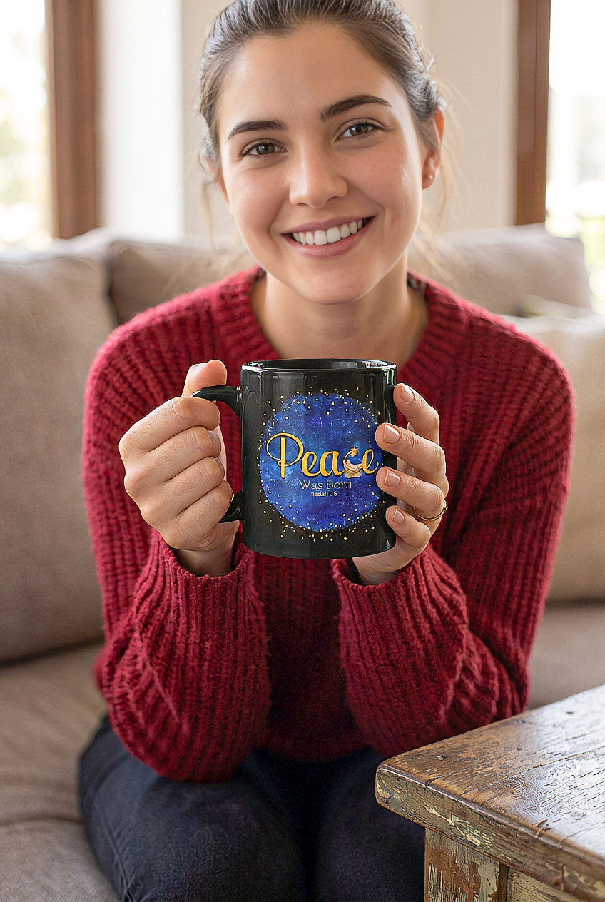 Smiling woman in a red sweater holding a black Peace Was Born Isaiah 9:6 mug while seated beside a wooden table