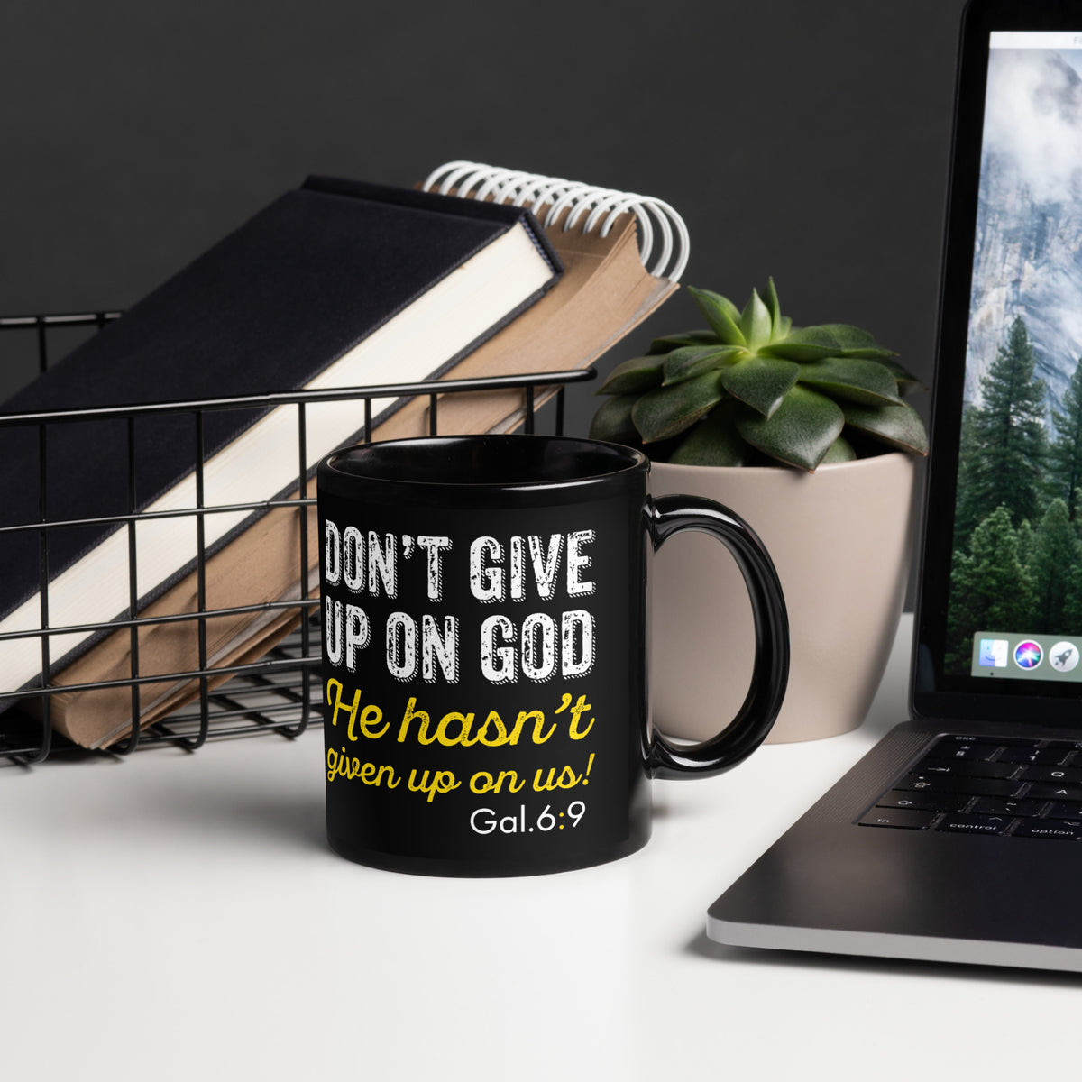 Black Don't Give Up on God 11 oz mug beside a laptop, notebooks and succulent, handle on right
