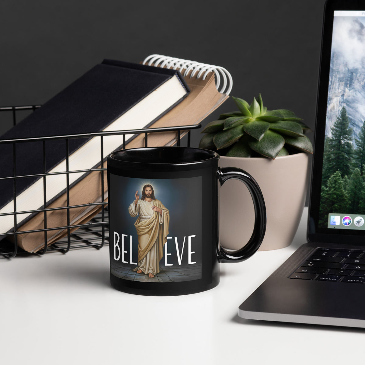 Black Believe In Jesus 11 oz mug beside a laptop, notebooks and succulent, handle on right