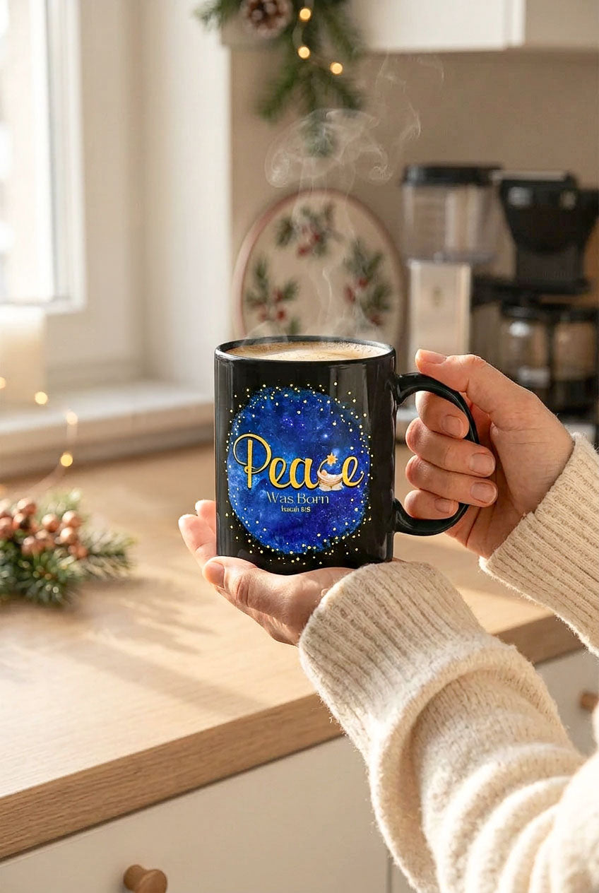 Hands in cream sweater sleeves holding a steaming black Peace Was Born mug in a kitchen decorated for Christmas, handle on the right