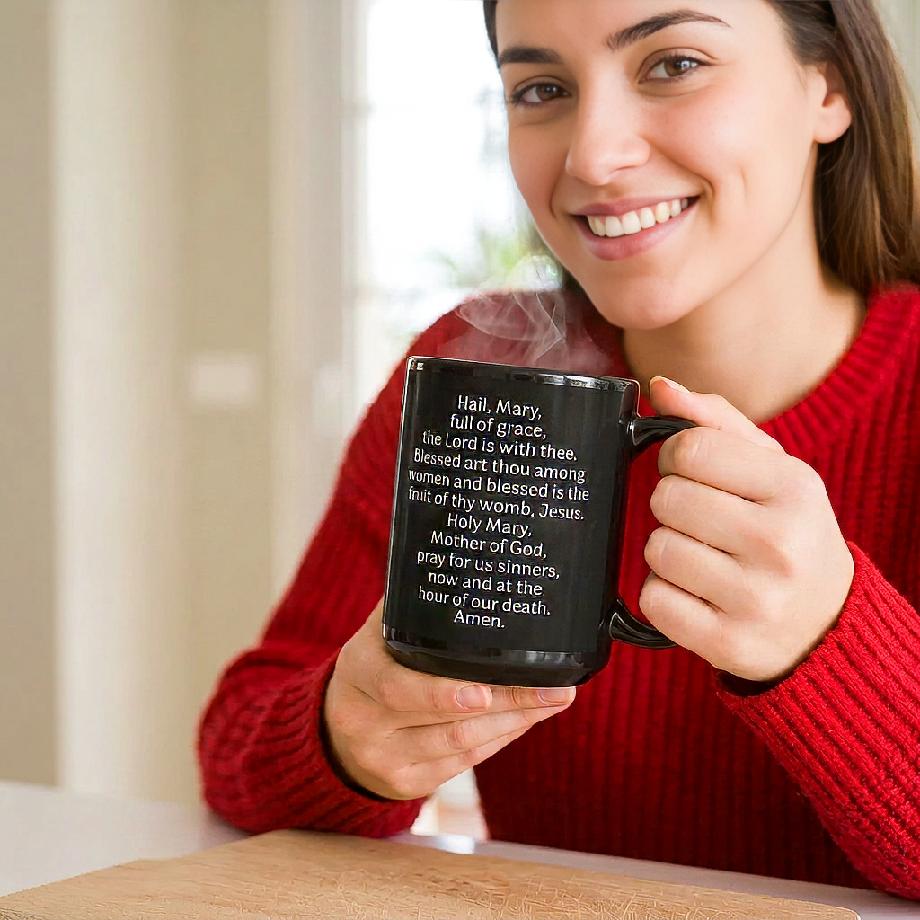 Smiling woman in a red sweater holding a steaming black mug displaying the Hail Mary prayer in white lettering