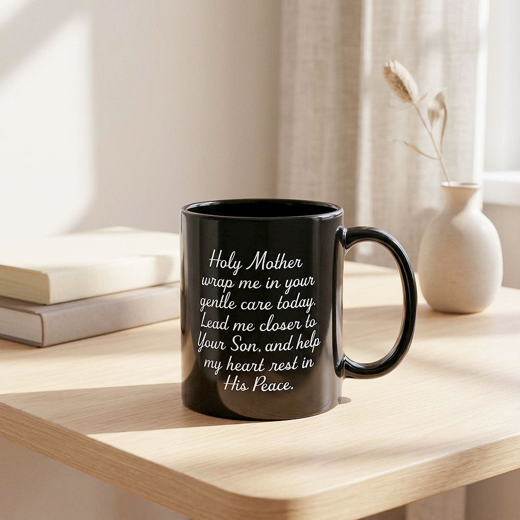 Black Holy Mother prayer mug on a sunlit table beside stacked books and a vase, handle on the right