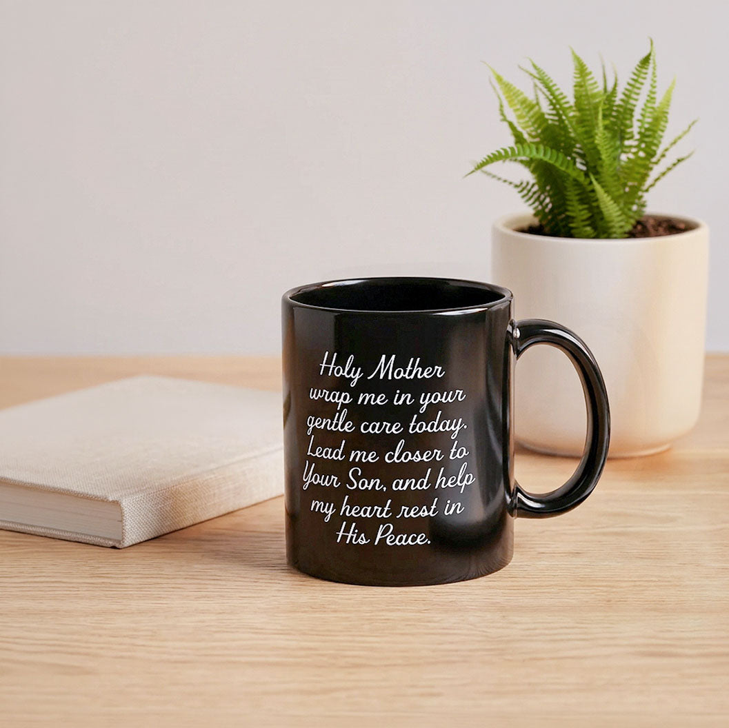 Black Holy Mother prayer mug on a wooden table beside a notebook and potted fern, handle on the right