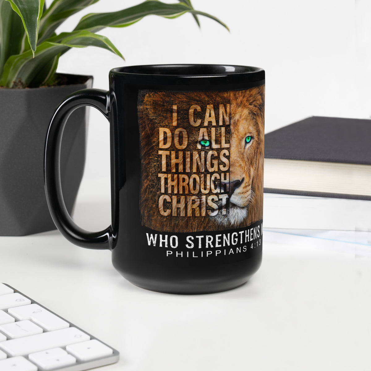 Black Philippians 4:13 lion mug beside a keyboard, plant and notebook on a white desk, handle on left