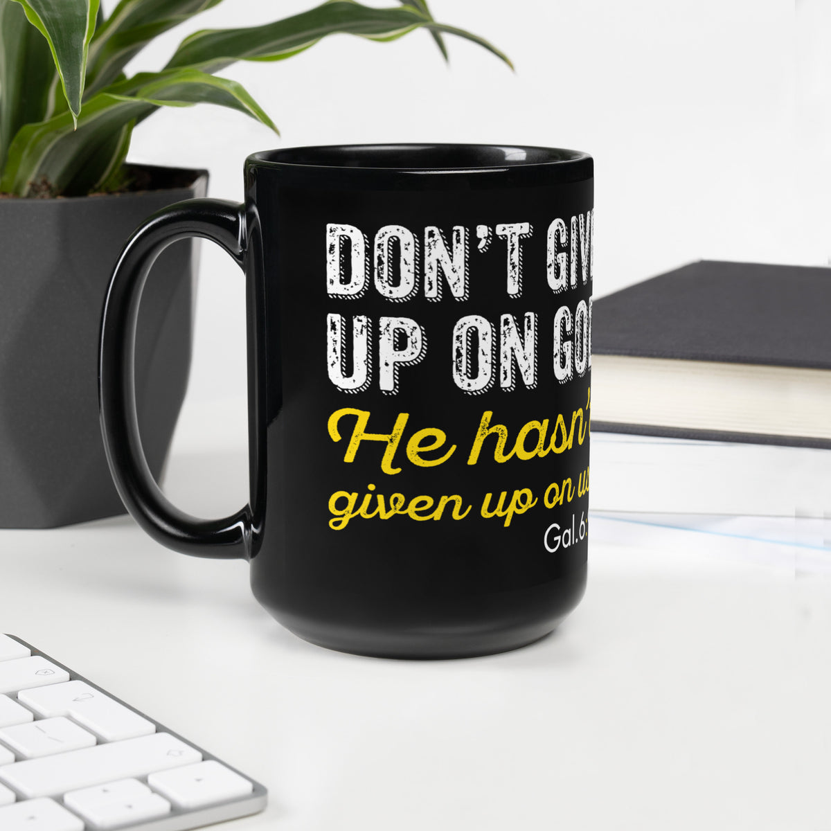 Black Galatians 6:9 15 oz mug on a white desk beside a keyboard, plant and notebook, handle on left