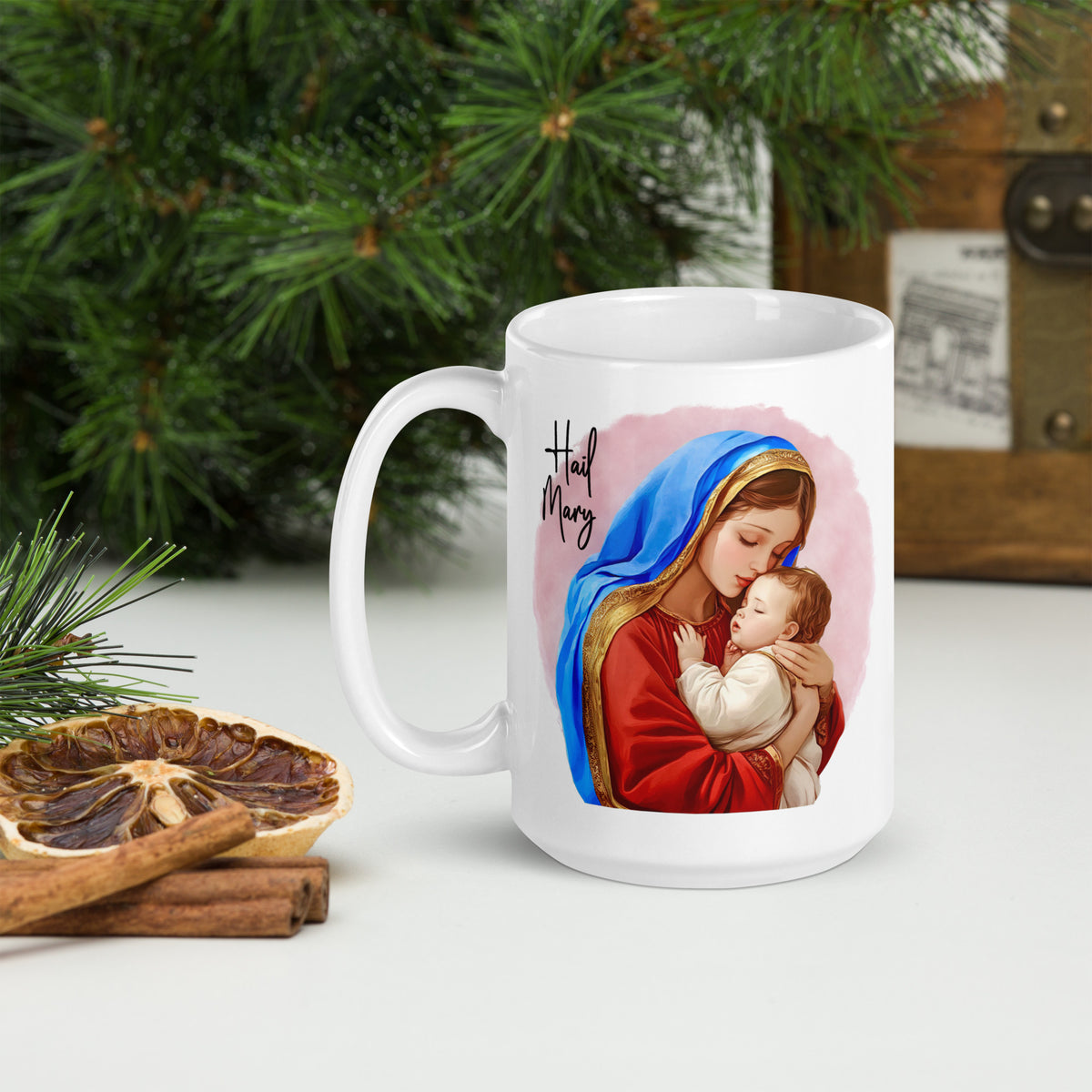 White Hail Mary mug beside evergreen branches, dried orange and cinnamon sticks, handle on the left
