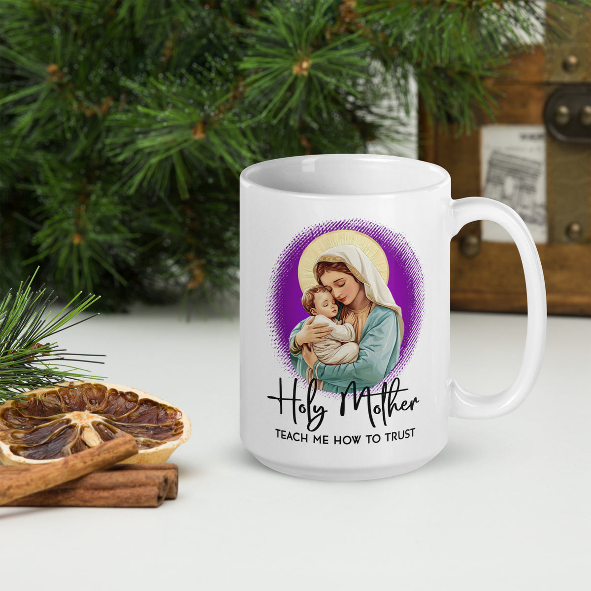 White Holy Mother Teach Me How to Trust mug beside evergreen branches, dried orange and cinnamon sticks, handle on the right