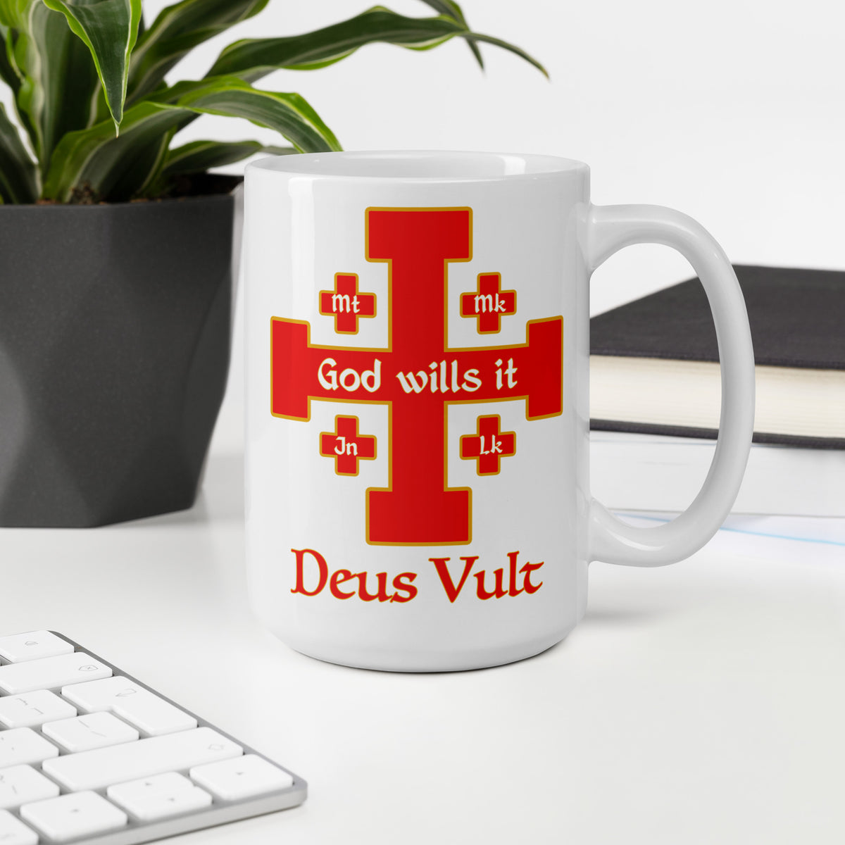 White Deus Vult cross mug on a desk beside a keyboard, plant and books