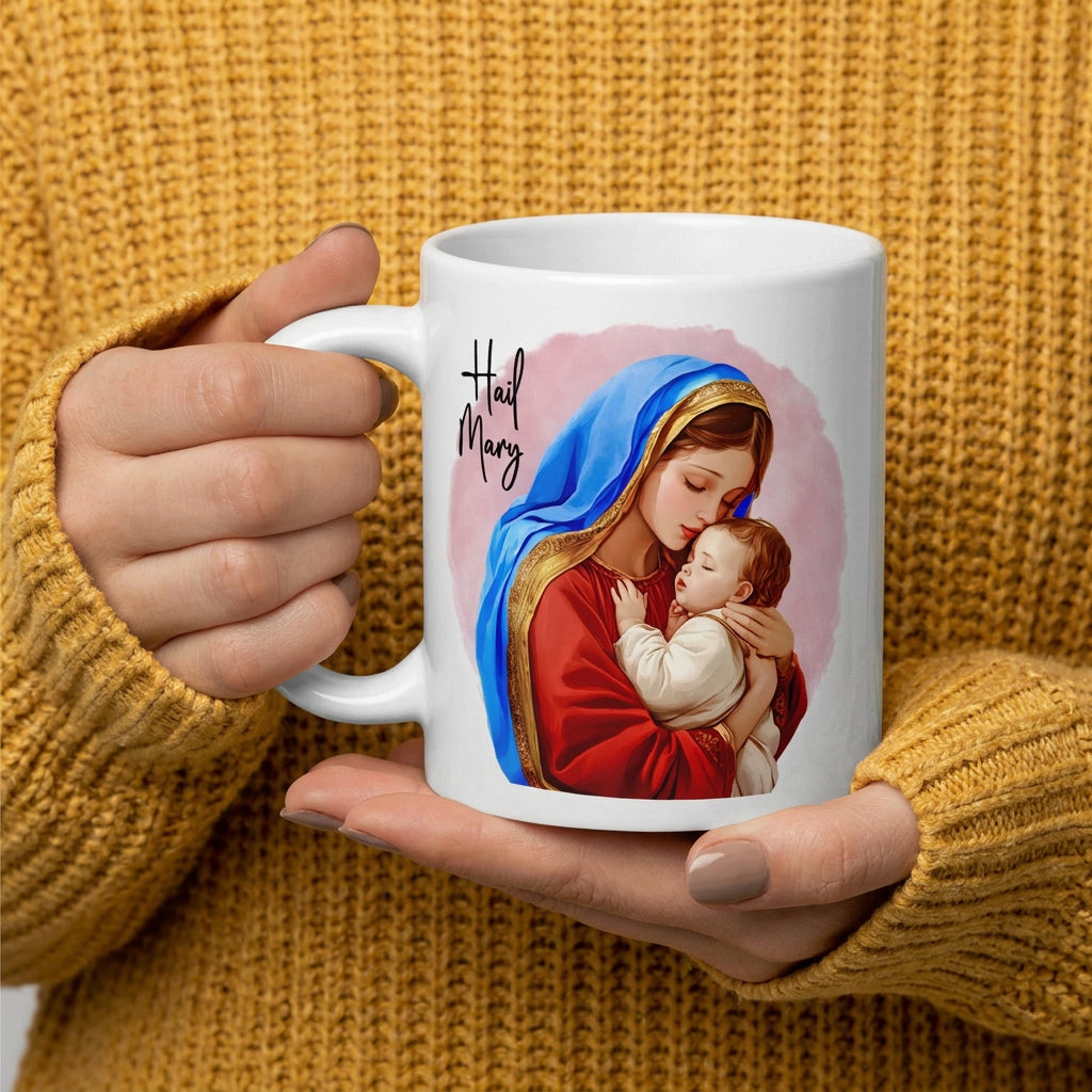 Person in a mustard knit sweater holding a white mug featuring Mary embracing Jesus and Hail Mary lettering